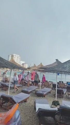 The video shows a beach with many empty lounge chairs and thatched umbrellas. A few people are walking on the beach. The sky is overcast and the water is calm.