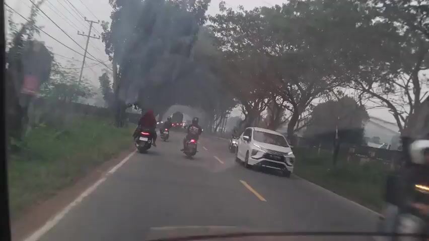 The video shows a point-of-view recording from inside a vehicle, looking forward through the windshield. It depicts a road with traffic, primarily motorcycles and cars. The road is paved with a yellow line marking the center. On the left side of the road, there is a grassy verge and some vegetation, with trees arching over the road in places, creating a canopy. On the right side, there are more trees, some power lines, and a few structures, including a small building or shed. Several motorcycles are visible, some with riders wearing helmets and jackets, moving in the same direction as the camera vehicle. A white car is also in frame, to the right of the road. There are hints of smoke or haze in the air, particularly in the distance. The lighting suggests it is daytime, possibly early morning or late afternoon, with overcast conditions.