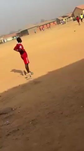 The video shows a dirt field where a group of people are playing soccer.  The field is surrounded by simple buildings.  A person in red soccer gear juggles a soccer ball.  Other players run across the field.  There are a mix of people in athletic attire.  The scene is bathed in sunlight.