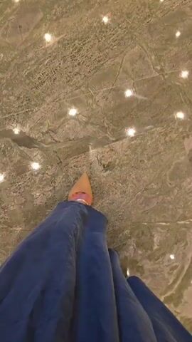This is a first-person point-of-view video of someone walking. The person is wearing a long, flowing blue dress and pointed yellow high heels. The shoes are visible at the bottom of the frame. The ground is made of marble tiles, with alternating sections of green and reddish-brown marble, separated by cream-colored marble strips. Small, bright lights are embedded in the floor at regular intervals, reflecting off the polished surface of the marble. The camera angle is looking down at the person's feet and the floor as they walk forward. The lighting appears to be dim, with the embedded lights providing the primary illumination.