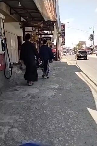 The video begins with a view of a street, possibly in Southeast Asia, with buildings on either side. A black SUV is seen driving away from the camera on the road. The camera then shifts to the perspective of someone on a motorcycle, showing a rearview mirror. The mirror reflects a bright blue sky and the faces of two people wearing white hijabs. As the video progresses, the motorcycle appears to be moving, and the reflection in the mirror shows more people and a building with an archway. The street is paved, and there are power lines visible. The sun is shining, casting shadows on the road. The environment appears to be a public street with businesses and residential buildings.