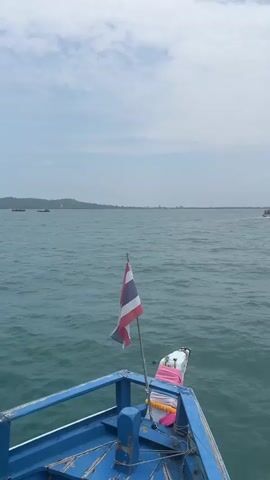 The video captures a boat journey across the waters of Ao Wiang Wan, Thailand. The scene opens with a view from the bow of a blue, longtail-style boat, featuring a fluttering Thai flag and ceremonial cloths draped on its front. The camera pans from left to right, revealing the expansive, choppy teal-colored sea under an overcast sky.

In the distance, a hazy coastline with rolling mountains is visible on the horizon. Another boat can be briefly seen in the early part of the video, also traveling across the water. The weather appears to be overcast, but it is daytime, likely late morning or afternoon. No specific people are visible, but the perspective is from aboard the moving boat, suggesting passengers. No visible text is present. The overall atmosphere conveys a sense of serene travel on the open water.