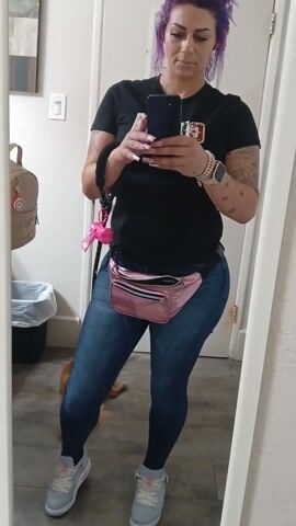 A person with purple hair stands indoors, taking a mirror selfie. They wear a black t-shirt with a graphic, blue jeans, and gray and white sneakers. A shiny pink fanny pack is around their waist. They hold a dark-colored phone in their hands, wearing an Apple Watch and bracelets on their left wrist. Tattoos are visible on their arms. A dog lies on the floor near the bottom of the frame. A backpack hangs on the wall to the left of the mirror, and a trash can is visible behind it. A light switch is on the wall above the backpack. A white door is to the right of the mirror.