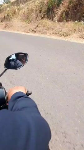 The video shows a first-person perspective of someone riding a motorcycle on a paved road. The road is winding and appears to be in a rural or hilly area, with dry vegetation and scattered trees on either side. The sky is blue with some white clouds. There is a traffic sign visible that appears to prohibit U-turns. The rider's hand is visible on the handlebars, and a portion of the motorcycle's mirror is in the frame. The overall impression is a scenic ride through a dry, possibly arid landscape.