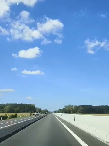 A point-of-view shot from inside a vehicle driving on a multi-lane highway. The road is paved and marked with white lines. On the left side of the road is a barrier and a field with trees in the distance. On the right side of the road is a concrete barrier. The sky is bright blue with scattered white clouds. Several vehicles are visible in the distance on the highway, traveling in the same direction as the camera vehicle. The overall scene suggests travel on a sunny day.