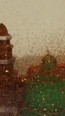 This is a close-up video of a miniature model. On the left side of the frame is a brown tower-like structure with silver bands. To the right of the tower is a green dome-shaped structure, also with some brown glitter. Between the two structures, and scattered across the entire scene, are numerous small, dark specks and some sparkles. The background is a light, solid color. The camera appears to be slightly moving, or there's a subtle vibration causing the scene to shift slightly.