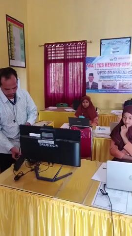 A person is standing and working on a computer setup in a room. The room appears to be a classroom or office. There are several students, all wearing hijabs, seated at desks working on laptops. A banner in the background reads "TKA (TES KEMAMPUAN AKADEMIK)" which translates to "Academic Ability Test." The desks are covered with yellow tablecloths.  The person standing is wearing a light-colored patterned shirt and has a microphone attached to their shirt. The computer monitor in front of them has a label that reads "DAU/02/2024."