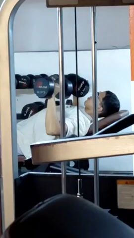 A man is doing dumbbell bench presses at a gym. He is lying on an inclined bench with his feet on the floor. He is wearing a white t-shirt and glasses, and he is holding a dumbbell in each hand. He raises the dumbbells over his chest, pauses, and lowers them. In the background, there are shelves of dumbbells. The camera is stationary and is angled towards the man. The foreground is partially blocked by a weight machine.
