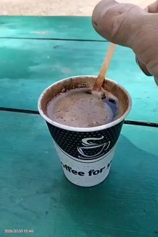 A close-up, over-the-shoulder shot shows a hand stirring a cup of coffee with a wooden stick. The coffee is in a white paper cup with a black dotted pattern and the words "coffee for you" visible. The coffee is dark brown and frothy. The wooden stick is being moved in a circular motion, creating ripples on the surface of the coffee. The background is out of focus, but appears to be a green wooden surface. The timestamp in the bottom left corner reads "2026.07.05 13:40".