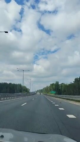 A point of view shot from a vehicle traveling on a highway. The road is asphalt with white and yellow lane markings. On both sides of the road, there are metal guardrails and dense green trees. Tall streetlights are visible along the left side of the highway. The sky is bright blue with scattered white, fluffy clouds. There are other vehicles on the road, including a dark-colored SUV on the left lane passing the camera's vehicle. A green highway sign is visible in the distance on the right side of the road. The overall scene depicts a daytime drive through a forested area on a highway.
