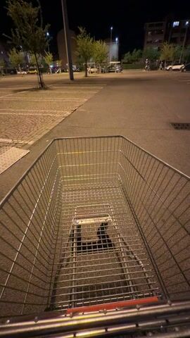 The video shows the first-person perspective of someone pushing an empty shopping cart across a paved surface at night. The cart is metal with a wire mesh bottom. The paved surface appears to be a parking lot or street with streetlights and the blurred lights of cars in the background. There are buildings and trees visible in the distance. A person walks past the cart. The focus is primarily on the interior of the cart and the immediate surroundings.