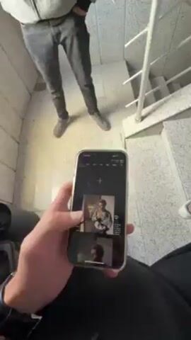 A person wearing black pants and holding an iPhone with a gold trim is in a stairwell. They are pointing the phone down the stairs and taking a photo. In the background, another person wearing gray pants and a light gray sweatshirt is standing still with their hand in their pocket. The stairwell has a gray, textured floor and white railings. The camera app is open on the phone, showing the live view.
