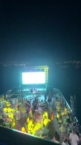 The video displays a night scene on the deck of a boat. The deck is crowded with people seated at tables and standing. A large screen displaying the text "OCEAN VISION" is visible at the front of the deck, illuminated with changing colored lights, predominantly blue and pink. There are speakers and lighting equipment around the screen. The boat appears to be on water, with city lights visible in the distance across the water, indicating it is near land. The overall atmosphere suggests a party or entertainment event on the boat.