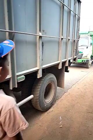 The video opens with a close-up of a young boy wearing a blue and orange baseball cap and a tan t-shirt, seen from behind as he looks towards a large gray truck. The scene then pans to the right, revealing a green and yellow truck with "WATJOLAR" written on its side. The truck has a large yellow tank attached to its trailer. Another person, a woman wearing a maroon shirt and yellow pants, walks past the camera. The camera continues to focus on the green and yellow truck, showing its front and side. A young child's head is visible at the bottom right of the frame. The video captures the truck parked on a dirt road, with a gray truck in the foreground. The environment appears to be outdoors, possibly in a rural or semi-urban setting, with power lines visible in the background.