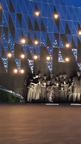 Here's a concise description of the video: 

The video shows a memorial featuring a group of bronze statues of people holding what appear to be lanterns or candles. Above the statues is a wall decorated with geometric shapes, possibly triangles or chevrons, with lights interspersed among them. The scene is set at dusk or night, with a blue sky visible in the background. The area in front of the memorial appears to be a paved plaza.