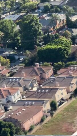 The video shows an aerial view of a suburban area. Buildings with tile roofs are visible, interspersed with trees. Some buildings appear to be apartment complexes, while others look like single-family homes. A few cars are parked on the street and in driveways. In the foreground, a large industrial building with a white roof is visible, and further in the distance, the landscape stretches out to rolling hills under a clear blue sky. The sun is shining, casting shadows.