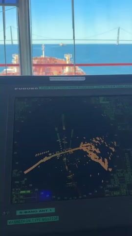 A close-up shot shows a radar screen on a ship's bridge. The radar displays a circular display with various blips and lines, indicating targets and navigational information. Above the radar screen, a window reveals a view of the sea, with a large red and white ship visible in the foreground and a distant bridge in the background. The sky is clear and blue. The overall scene suggests a maritime environment, likely the control center of a vessel at sea.