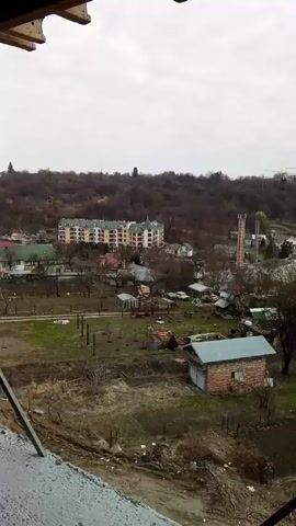 From a high vantage point, the video captures a cityscape under an overcast sky. The foreground shows a construction site with exposed dirt and construction materials. Buildings of varying heights and styles dot the landscape, including apartment complexes and individual houses. Trees are interspersed throughout the city, most without leaves, suggesting it is autumn or winter. A crane can be seen on the horizon, indicating ongoing construction. The camera moves slightly, showing different parts of the city. No people are visible in the frame.