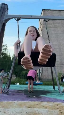 This video captures a playful scene at a playground in Boksitogorsk, Russia. The primary focus is on a young girl on a swing, captured from various angles as she moves back and forth. She is wearing a white tank top, denim shorts, and pink sneakers. Her hair is styled in a way that shows it blowing in the wind as she swings. In the background, other children and adults are present on the playground. One child is on another swing set, and another child appears to be on a scooter. The playground has a multi-colored surface, with patches of purple, green, and beige. Behind the playground, there's a tall, brick apartment building and some trees. The sky is visible and appears to be partly cloudy, suggesting it might be late afternoon or early evening. The overall mood is cheerful and active, indicative of a typical day at a public park.