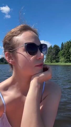A young woman with blonde hair tied back and wearing sunglasses and a striped bikini top is sitting on a paddleboard in a lake. She rests her chin on her hand and looks to her right. The paddleboard is orange and white with some text on it. The water is dark and rippling, and the sky is bright blue with a few white clouds. Trees and a hill are visible in the background.