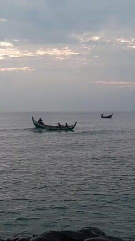 The video shows two small fishing boats with people on board in a calm sea under a cloudy sky. The foreground boat has several people in it.  The background boat has only one person visible. The boats appear to be moving away from the shore.  The water is relatively calm with small waves.  The light suggests it could be early morning or late afternoon.