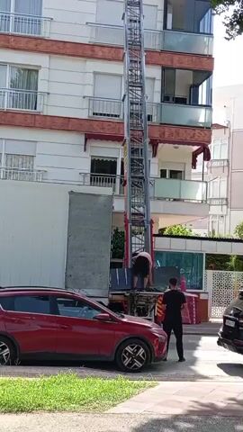 The video shows a moving operation in an urban environment. A tall metal lift with a platform is attached to the side of a moving truck, extending upwards towards a multi-story apartment building. Two individuals are visible near the base of the lift, appearing to be moving items. One person, wearing a dark shirt and pants, stands facing the lift, possibly directing or observing. Another person, wearing a light-colored shirt and dark pants, is positioned on the lift's platform, bent over and handling objects. The moving truck is white and has its rear doors open, revealing its interior, likely filled with furniture or belongings. A red car is parked to the left of the truck, and another vehicle, partially visible on the right, suggests a street or parking area. The apartment building has balconies and windows, with some awnings visible. Greenery, including trees or bushes, is present along the sidewalk. The overall scene depicts a coordinated effort to transport items to or from an upper floor of the building.