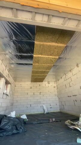 The video shows the interior of a room under construction. The walls are made of white cinder blocks, and the floor is covered with black plastic sheeting. The ceiling is being insulated with yellow fiberglass material and covered with a silver reflective vapor barrier. Some black plastic sheeting is also visible on the ceiling. There are stacks of what appear to be building materials, including yellow insulation and rolls of silver material, on the floor. The overall impression is of a space being prepared for finishing.