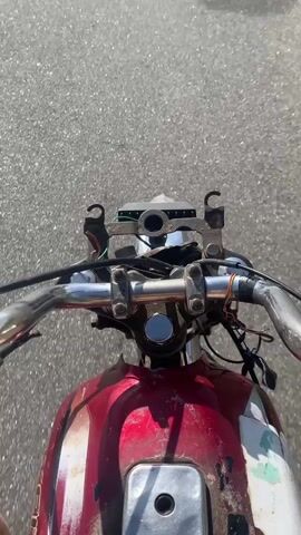 This video captures a first-person perspective from a motorcycle in Nigeria. The motorcycle appears older, with a red and white painted fuel tank and exposed wiring near the handlebars. The dashboard area is rudimentary, with visible metal components and some greenery peeking out. The video begins on a paved road, the asphalt clearly visible, and the motorcycle seems to be in motion.

As the video progresses, the scenery shifts. The paved road gives way to a rougher, gravelly track. To the right of the motorcycle, the landscape opens up to a dry, semi-arid environment. There are scattered acacia-like trees with sparse foliage, typical of northern Nigerian vegetation. The ground is a mix of sand and small stones. The sky is clear and bright, suggesting a sunny day, likely mid-morning or early afternoon given the intensity of the light and the shadows.

The overall mood is one of exploration or travel through a rural setting. There are no people visible in the footage, emphasizing the solitary journey. The activity is simply riding the motorcycle through different terrains. The rough texture of the road and the dry vegetation are the most notable environmental details. There is no visible text in the video.