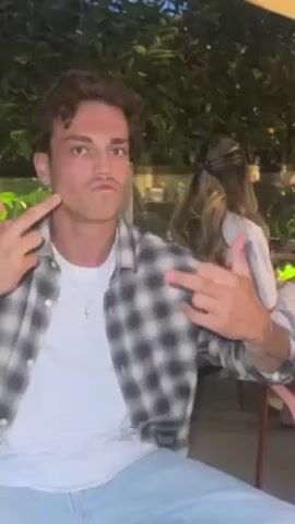 A young man with curly brown hair, wearing a white t-shirt, light blue jeans, and an open gray and white plaid shirt, sits outside making a funny face and holding up two fingers on each hand. A woman with long brown hair sits behind him wearing a white shirt and her back is to the camera. There are green plants behind them.
