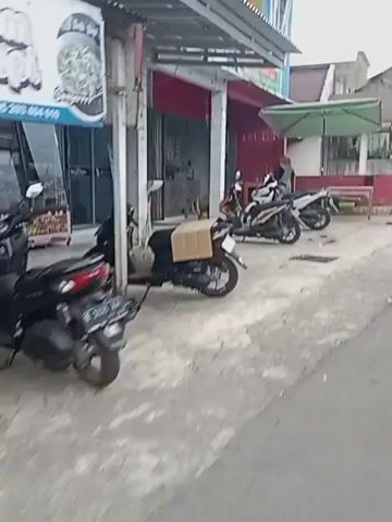 Two motorcycles are parked on a sidewalk next to a street. The street appears to be paved and cracked. On the sidewalk, there is a table with a green umbrella over it. Further down the street, there is a building with red accents. There are people walking and sitting on the sidewalk. The environment appears to be an urban or suburban area.