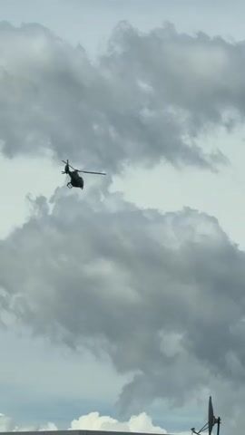 The video shows a helicopter flying against a cloudy sky. The helicopter is dark in color, and its blades are rotating. The clouds are gray and white, and they fill the frame. The camera is pointed up at the helicopter. There is a structure with an antenna visible in the lower right of the frame.