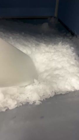 The video shows a person using a white plastic ice scraper to break up ice in a bin. The ice is finely chopped and white, filling the bin. The interior of the bin appears to be blue. The scraper is moved across the ice surface.