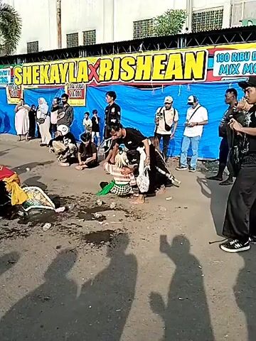 The video captures an outdoor cultural event or performance on a sunny day in Sragen, Indonesia.

**Subjects**: Numerous people are present, including several young men performing a dynamic, possibly ritualistic, dance. These performers wear varied attire, including striped shirts, dark pants, and head coverings. Some are seen in crouching or lying positions on the ground. A large crowd of spectators, dressed in casual clothing, gathers around, observing the performance; some are recording it with their phones.

**Objects**: The scene features several temporary market-style tents with colorful awnings. A small truck or cart equipped with drums serves as part of the performance setup. Motorcycles are parked among the spectators. The ground directly around the performers is wet and contains scattered debris, possibly offerings or trash. Banners are visible in the background.

**Scene/Setting**: The event takes place on a paved outdoor street or open area, framed by buildings and palm trees in the background. The setup with tents and a crowd suggests a festival, market, or public celebration.

**Activity or event taking place**: The central activity is a traditional performance or parade. The performers engage in energetic movements, with some individuals appearing to be in a trance-like state as they interact with the ground or other performers. Spectators are actively watching the proceedings.

**Notable details**: The time of day is clear, indicated by bright sunlight and a blue sky, suggesting fair weather. The atmosphere appears lively and engaging.

**Visible text**: A large blue banner in the background displays "SHEKAYLA X RISHE...". Another white banner, partially visible, reads "KIRAP..." and "TEM...", likely part of a phrase such as "Kirab Budaya" (Cultural Parade). The word "JEMAS" can be seen on one of the tents.