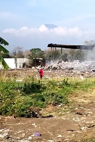 A wide shot captures an outdoor scene with a person in a red shirt walking through a cluttered area filled with debris. In the background, a mountain is visible against a sky with scattered clouds. To the left, banana leaves hang down, and a white structure is partially visible. In the center and right, there is a large pile of trash, with some smoke rising from it, suggesting a fire or burning activity. A rustic structure with a dark roof stands behind the trash heap. The ground in the foreground is muddy and littered with various types of trash. The overall environment appears to be a makeshift waste disposal or recycling area.
