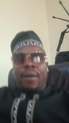 A man is in an office. He is wearing sunglasses and a black hat. He is wearing a black shirt with a white pattern on it. He is sitting in a black chair. He is chilling and trying to do some work.