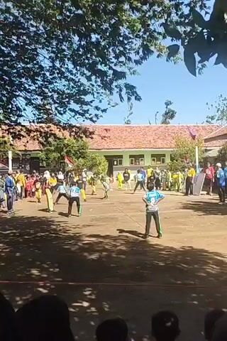 A group of children are playing a game of volleyball on a dirt court in front of a school building. The children are wearing brightly colored athletic clothing. The school building has a red tile roof and is painted green. There are trees and foliage surrounding the court, and the sky is clear and blue. A crowd of spectators is watching the game from the sidelines. The game appears to be in progress, with several children actively engaged in playing.