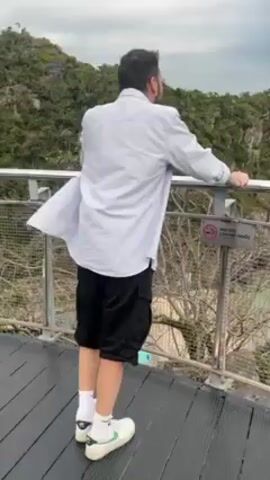 The video shows a man standing on an observation deck overlooking a scenic ocean view. He is wearing a light-colored, long-sleeved shirt, black shorts, white socks, and cream-colored sneakers with green accents. He leans against a metal railing, gazing out at the water.  The camera then pans to show a wide view of the ocean, dotted with several small, rocky islands covered in vegetation. The sky is overcast. There is a sign with text in a foreign language and a symbol indicating no smoking on the railing.  The observation deck appears to be made of a metal grate.