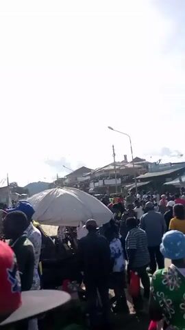 The video shows a large crowd of people gathered in what appears to be a bustling marketplace or town center. The camera moves through the crowd, capturing various individuals of different ages and wearing diverse clothing. Some people are carrying items, possibly goods for sale or personal belongings. The environment includes buildings, streetlights, and overhead wires, suggesting an urban or semi-urban setting. The sun is shining brightly, creating a somewhat overexposed visual. The scene conveys a sense of activity and public gathering.