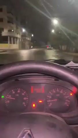 A video shows a view from inside a car driving at night in what appears to be a city. The camera focuses on the car's dashboard, which displays various gauges and warning lights. The speedometer reads approximately 20 kilometers per hour. Through the windshield, the wet road is visible, reflecting streetlights. Buildings line the street, and another car is seen in the distance ahead. The steering wheel is in the foreground, and a piece of paper rests on the dashboard.