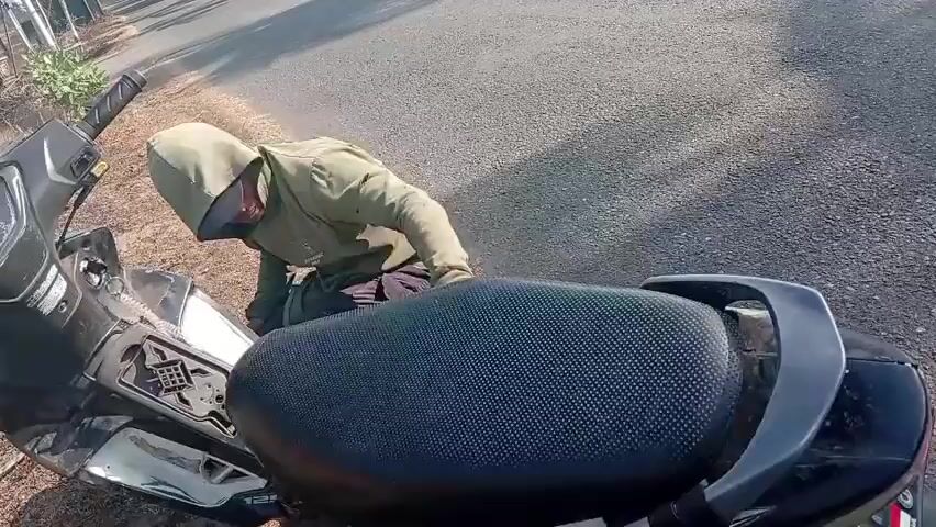 A person wearing a hooded jacket and a mask is kneeling next to a motorcycle on the side of a paved road. The motorcycle is black and appears to be a scooter. The person is reaching towards the rear of the motorcycle. The environment is outdoors with trees lining the side of the road. The sun is shining, casting shadows.