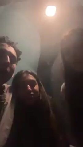 The video features three young adults seemingly taking a selfie indoors, under a bright overhead light. One person on the left is a young man with dark curly hair, wearing a light-colored shirt, and smiling. The person in the middle is a young woman with long, dark, wavy hair. She is facing the camera with a neutral expression. The person on the right is another young woman with long, dark hair, partially obscured by the lighting. The background appears to be a room with a light blue or green wall. The lighting is bright, casting shadows on the subjects' faces.