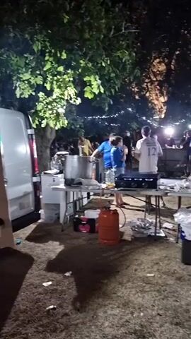 The video shows a nighttime outdoor gathering, likely a festival or event, with people milling around. In the foreground, there are several folding tables set up. On these tables are cooking equipment, including gas burners and what appears to be a large pot. Two large orange propane tanks are visible. In front of the tables, there is a grassy area with scattered trash, including cardboard boxes and plastic bags. Several people are standing near the tables, some appearing to be involved in food preparation or serving. In the background, there are more people, tents, string lights, and bright spotlights, illuminating the scene. The overall atmosphere seems to be a bustling outdoor event at night.