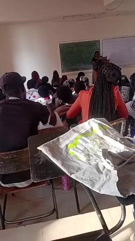 The video shows a classroom setting with a teacher standing at a lectern facing a group of students seated at desks. The students are mostly seen from the back, and some appear to be taking notes. There is a green chalkboard and a whiteboard behind the teacher, both with writing on them. The classroom is well-lit, and the desks and chairs are a mix of wood and metal. A gray shirt with a green design is lying on a desk in the foreground.