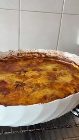 The video shows a close-up shot of a freshly baked dish in a white, ruffled baking dish, sitting on a wire rack inside an oven. The dish appears to be a baked casserole or moussaka, with a browned top layer that has a slightly glossy sheen, suggesting the presence of cheese and sauce. The edges of the dish are also browned, indicating they have been exposed to heat. The background features white tiles, providing a clean and neutral backdrop.