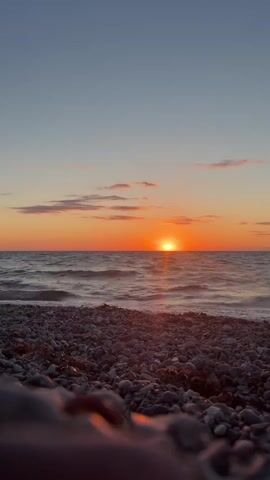 The video shows a low-angle view of a beach at sunset. The foreground is filled with smooth, grey and brown pebbles, some of which are wet and glistening. The ocean stretches out behind the pebbles, with gentle waves lapping at the shore. The sun is setting on the horizon, casting a warm, orange glow across the sky and water. There are a few wispy clouds in the sky, tinged with pink and orange. The sky transitions from a deep blue at the top to a lighter orange near the horizon. There are no people or man-made objects visible in the video. The overall atmosphere is peaceful and serene.