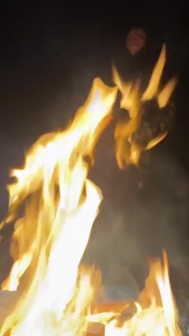 The video shows a person standing in front of a bonfire at night. The person is wearing a camouflage jacket, dark pants, and boots. They have their hands in their pockets. The bonfire is large and bright, and smoke is rising from it. The background is dark.