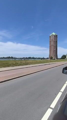 A water tower with a green roof and a reddish-brown brick structure is visible on the right side of the frame. It is situated near a body of water. A road runs in the foreground, with a sidewalk or pedestrian path adjacent to it. The sky is blue with some white clouds, indicating a sunny day. The camera appears to be in motion, possibly from inside a vehicle, as the side mirror of a car is visible in the lower right corner. Trees and vegetation are present in the background.