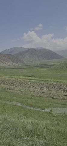 A slow panning shot of a mountain valley on a cloudy day. In the foreground, a grassy hill slopes down towards a patch of tilled earth. Further back, a wide, green valley stretches out, dotted with patches of yellow flowers. In the distance, rolling hills give way to rugged, snow-capped mountains. The sky is a pale blue, with scattered white clouds. The overall impression is one of vastness and natural beauty.