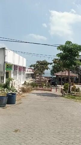 The video pans across an outdoor scene, likely a boardwalk or a recreational area. In the foreground, a paved walkway made of interlocking bricks leads towards a cluster of low-rise buildings and a gazebo. To the left, a white building with several closed storefronts is visible, with a blue trash bin in front of it. Several trees and picnic tables are scattered throughout the area, with some string lights draped between poles. A motorcycle is parked near the gazebo. In the background, a body of water, possibly the sea, is visible, along with a clear sky with scattered clouds. A larger building with a sign is situated behind the cluster of smaller structures. The scene appears calm and empty.