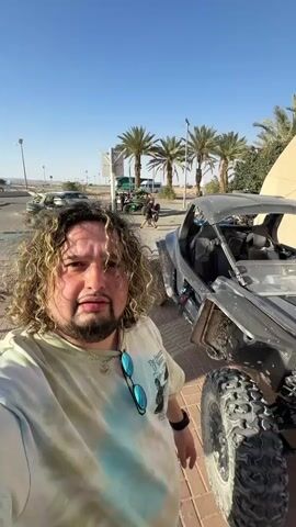 Here's a detailed description of the scene.

A man with curly blond hair and a beard stands in front of a gray off-road vehicle. He is wearing a tie-dye t-shirt and sunglasses are hanging from his shirt. The vehicle is parked on a brick pavement. In the background, there are palm trees, other off-road vehicles, and people. The sky is clear and blue. The man is looking at the camera.